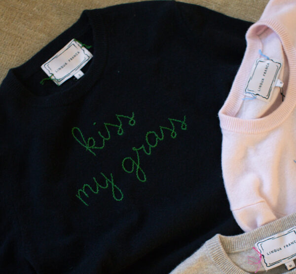 A Lingua Franca x Perfect Earth Project collaboration featuring a black cashmere sweater with green embroidery that says 'Kiss My Grass.'