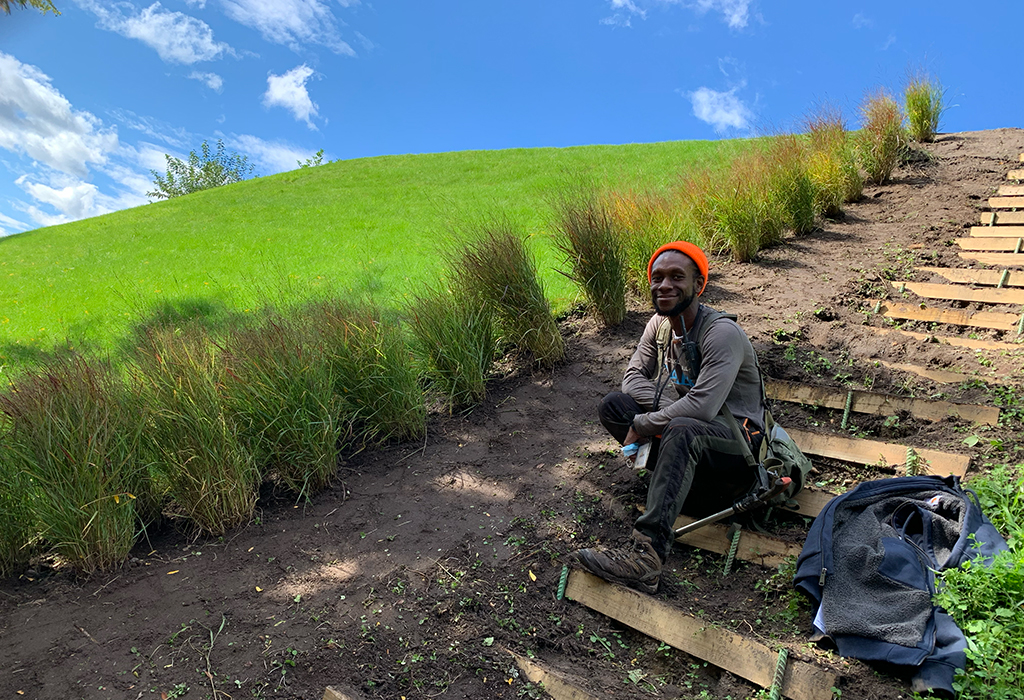 Rashid Poulson wearing a bright orange hat smiles while siting on the steps at one of the sound berms at Brooklyn Bridge Park.